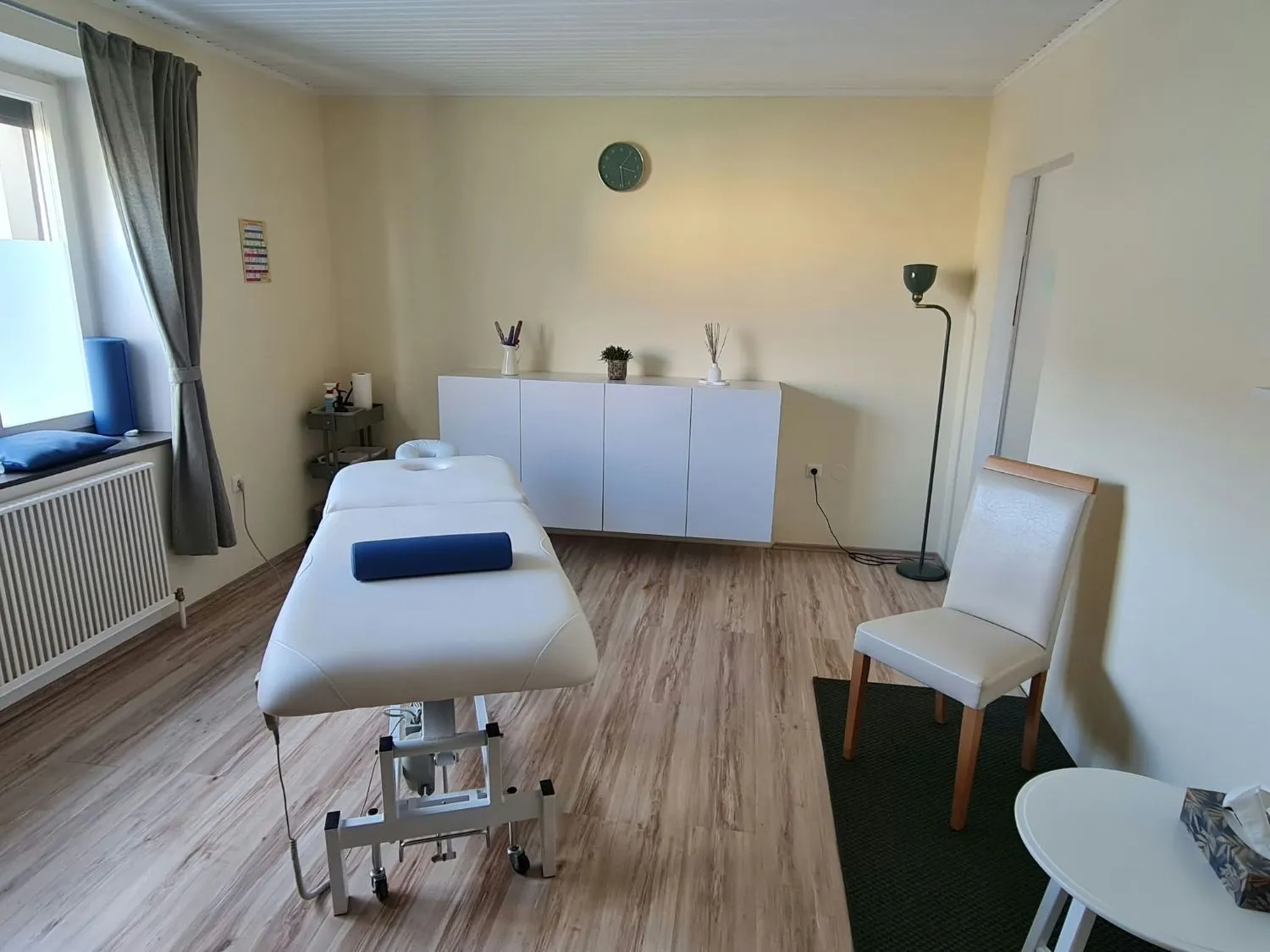 Behandlungsraum mit elektrischer Massageliege, Sideboard, Sessel und Stehlampe